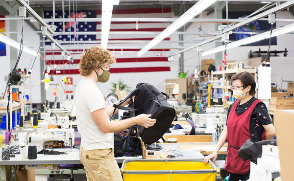 Nik holding a prototype of the Shadow Guide 33 backpack in our Seattle factory. He is talking with Fong and both are wearing masks. Rainbow, U.S.A. and Earth flags are in the background.